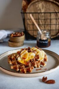 A round waffle on a ceramic plate topped with banana slices, chopped pecans, and a drizzle of caramel sauce, with a small jar of caramel and wooden cutting boards softly blurred in the background.