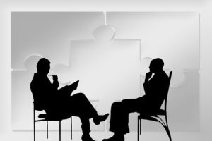 Two men in shadow seated across from each other in thinking poses, possibly considering a business writing coach.