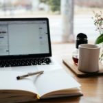 Pen sitting atop an open notebook in front of a laptop next to a mug on a table ready for a newsletter writer.