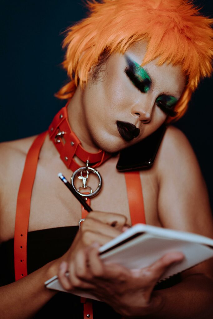 A man dressed as a female punk wearing an bright orange wig, green eyeshadow, and dark lipstick writing with a pencil on a pad of paper.