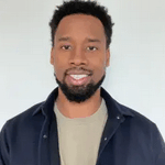 Brenton Thomas with a black beard, mustache, and hair smiling while wearing a dark jacket over a tan shirt.