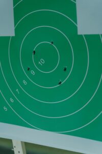 A green shooting target with concentric circles numbered 5 through 10, showing several bullet holes clustered near the center rings. The target is mounted on what appears to be a white stand or frame. It conceptualizes what can happen when you jot thoughts at random rather than organizing them clearly.