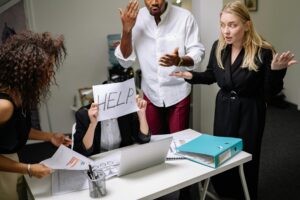 Four colleagues around a desk during a crisis communication strategy moment: a seated person holds a handwritten “HELP.” sign while others stand and gesture at a laptop.